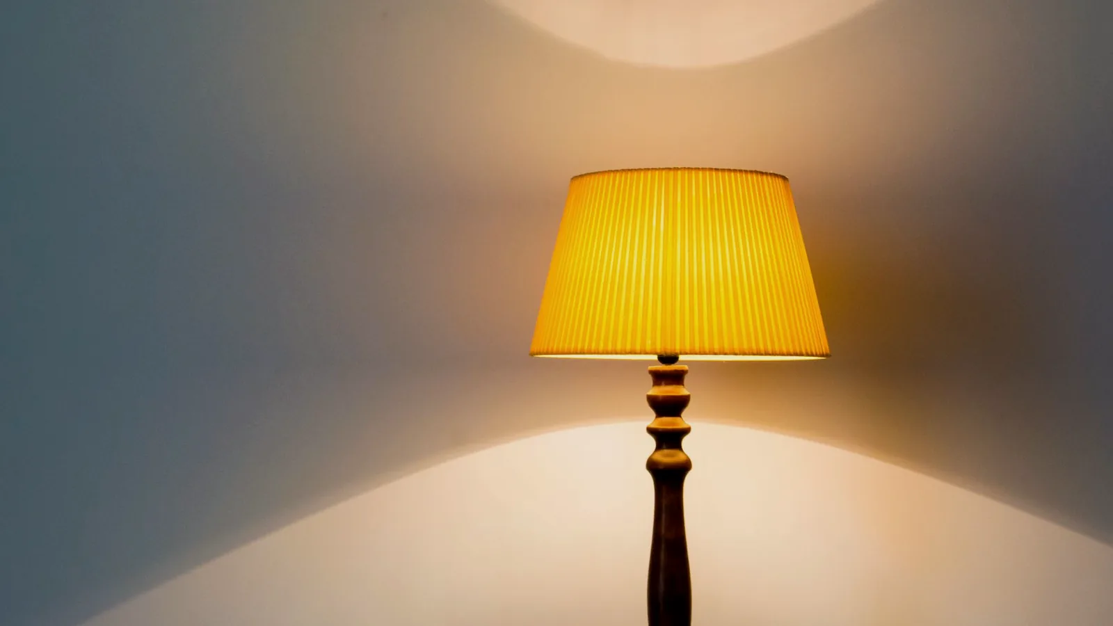 A pleated yellow lampshade glowing on a wooden floor lamp against a pale wall