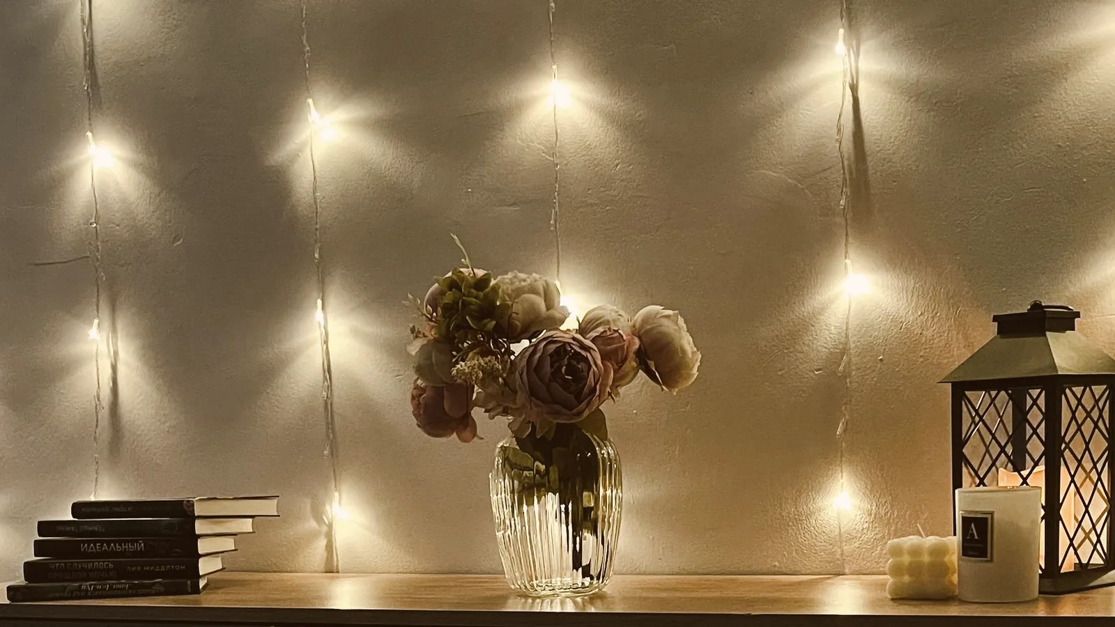 Warm fairy lights draped on a wall above a wooden console with flowers, a lantern and a wrapped gift